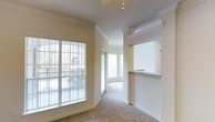 This single-floor residence offers a welcoming and open layout characterized by elegant architectural details and generous natural light. The living area is finished with soft neutral carpeting and warm beige walls, creating a calm and cohesive atmosphere throughout. Crown molding runs along the ceilings, adding a refined touch to the interior design.

Large windows and a sliding glass door line the exterior wall of the living space, flooding the room with natural light while offering views of an outdoor balcony area and surrounding architecture. The windows are fitted with vertical blinds, providing flexible light control.

A defining feature of the space is the pass-through window cut into the wall between the kitchen and the living area, complete with a countertop ledge — ideal for casual dining or serving. This architectural detail enhances the open feel while maintaining a functional separation between spaces.

The living area flows naturally toward an additional windowed alcove, providing a secondary area suitable for a dining nook or sitting space. The ceiling fan provides both comfort and practicality. Overall, the space presents a clean, classic interior with thoughtful design elements suited for comfortable everyday living.