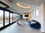This contemporary rooftop amenity space is designed with a clean, modern aesthetic that blends comfort and functionality. The centerpiece of the main lounge is a striking oval coved ceiling detail with warm ambient uplighting, adding architectural depth to the otherwise crisp white interior. Recessed lighting and large-format light-tiled flooring further enhance the bright, airy atmosphere throughout.

The lounge area is furnished with a deep navy blue sofa, accent chairs, and a marble-topped coffee table, arranged atop a patterned area rug to define the seating zone. Adjacent to this is a casual dining area featuring a round white table surrounded by vibrant red chairs, positioned near the built-in kitchenette. The kitchenette is outfitted with warm wood cabinetry, a wall-mounted flat-screen television, and under-counter appliances, making it well-suited for gatherings and events.

Floor-to-ceiling black-framed sliding glass doors span one full wall, flooding the interior with natural light and providing seamless access to the outdoor rooftop terrace, where open sky views await. Entry to the lounge is via a stairwell with sleek metal railings, and the transition area also features decorative wall art. The overall ambiance is polished and inviting, offering a sophisticated environment ideal for socializing or relaxing.