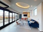 This contemporary rooftop amenity space is designed with a clean, modern aesthetic that blends comfort and functionality. The centerpiece of the main lounge is a striking oval coved ceiling detail with warm ambient uplighting, adding architectural depth to the otherwise crisp white interior. Recessed lighting and large-format light-tiled flooring further enhance the bright, airy atmosphere throughout.

The lounge area is furnished with a deep navy blue sofa, accent chairs, and a marble-topped coffee table, arranged atop a patterned area rug to define the seating zone. Adjacent to this is a casual dining area featuring a round white table surrounded by vibrant red chairs, positioned near the built-in kitchenette. The kitchenette is outfitted with warm wood cabinetry, a wall-mounted flat-screen television, and under-counter appliances, making it well-suited for gatherings and events.

Floor-to-ceiling black-framed sliding glass doors span one full wall, flooding the interior with natural light and providing seamless access to the outdoor rooftop terrace, where open sky views await. Entry to the lounge is via a stairwell with sleek metal railings, and the transition area also features decorative wall art. The overall ambiance is polished and inviting, offering a sophisticated environment ideal for socializing or relaxing.