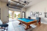 Located at 1633 Greenridge Dr in Burleson, TX, this single-room game and entertainment space is thoughtfully designed for social gatherings and recreation. The centerpiece of the room is a rustic solid-wood pool table with a bold blue felt surface, positioned atop a patterned area rug over rich wood-look tile flooring. Comfortable lounge chairs are arranged nearby, making for an inviting setting to relax between games. Along one wall, a built-in wet bar features dark cabinetry with granite countertops, an undermount sink, and a compact refrigerator — ideal for entertaining. A large wall-mounted flat-screen TV is positioned above the bar, adding a media element to the space. The walls are accented with curated gallery-style artwork, contributing warmth and personality to the room. A standout architectural feature is the full-width glass-panel retractable garage door that opens completely to a covered outdoor patio supported by brick and stone arched columns, seamlessly blending indoor and outdoor living. A ceiling fan overhead ensures comfort year-round. A glass-paned interior door provides access to an adjacent space, while the open-air connection to the landscaped exterior grounds enhances the overall sense of space and natural light throughout.