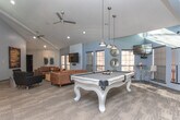 Located at 505 Riverbend Pkwy in Athens, Georgia, this single-floor community clubhouse is designed to offer a well-rounded social and functional environment. The space is defined by soaring vaulted ceilings that lend an airy, open feel throughout, complemented by abundant natural light streaming in through large windows along multiple walls.

The main lounge area is furnished with rich leather sofas and accent chairs arranged around a wood-and-granite coffee table, with a wall-mounted flat-screen television as a central focal point. Shiplap wall accents and contemporary artwork add texture and visual interest to the space.

Adjacent to the lounge, a dedicated billiards area features a striking white pool table beneath stylish pendant lighting, alongside a built-in kitchenette outfitted with cabinetry, countertop appliances, and a compact refreshment bar — ideal for casual gatherings.

A dining nook with built-in booth seating and pedestal tables sits beneath dramatic double-height windows, adorned with large-scale abstract artwork above, creating an intimate yet visually dynamic setting. Elegant pendant chandeliers illuminate this area with warm light.

A private conference room with glass-panel visibility is accessible from the lounge, providing a quiet workspace equipped with task chairs and a work table. The overall design blends modern, upscale aesthetics with comfortable, community-oriented functionality throughout.