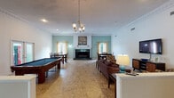 Located at 151 Century Dr in Greenville, SC, this single-floor facility offers a thoughtfully designed communal environment suited for wellness and recreation. The main gathering area is generously proportioned, featuring warm tile flooring, ornate crown molding, and soft teal accent walls that lend a calming, welcoming atmosphere. A traditional fireplace with dark tile surround and a white mantle serves as a striking focal point, flanked by dining tables and chairs ideal for group activities or meetings. A full-size billiards table anchors one side of the room, while plush leather seating arrangements provide comfortable lounging space throughout. Natural light flows in through sheer gold-toned drapery panels, adding warmth to the interior. An elegant chandelier and recessed lighting illuminate the space effectively. A corridor with rich hardwood flooring connects the main area to additional rooms, including a welcoming reception lounge with French doors opening to the outdoors, and a seating nook with blue accent chairs positioned outside a dedicated fitness room. The fitness room, accessible through glass-paneled French doors, features carpeted flooring and exercise equipment. Decorative crown molding is carried consistently throughout, unifying the space with a refined, residential character.