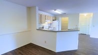 This single-story space offers a well-organized and functional floor plan designed for comfortable everyday living. The layout flows seamlessly from room to room, anchored by rich, dark hardwood-style flooring that runs throughout the main living areas, creating a warm and cohesive aesthetic.

The kitchen is a standout feature, designed with an open-concept peninsula that separates it from the living area while maintaining a connected, social atmosphere. It is equipped with light wood cabinetry providing ample storage, a white electric range, a refrigerator, and a double sink. The countertops offer generous prep space, and the peninsula serves as a natural gathering point.

The living area is open and airy, benefiting from natural light that filters in through windows, lending the space a bright and welcoming ambiance. The neutral white walls throughout provide a clean backdrop that complements the warm flooring tones.

Additional rooms visible in the floor plan include a bathroom fitted with standard fixtures, and supplementary spaces that offer flexibility for use as bedrooms or utility areas. The overall layout is practical and efficiently arranged, making thoughtful use of the available footprint. Clean lines, neutral finishes, and consistent flooring choices give the space a unified, move-in-ready character.