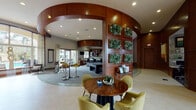This single-story clubhouse is designed with a contemporary, resort-inspired aesthetic that blends warm wood tones, lush greenery, and modern furnishings throughout. The entry foyer makes an immediate impression with soaring ceilings, rich wood-paneled walls accented by glass inlays, and a decorative mosaic tile floor medallion. A stylish reception console with geometric mirrored detailing anchors the space, flanked by natural light pouring through oversized glass doors that frame views of palm trees and a pool beyond.

The central gathering area is defined by a sweeping curved ceiling detail with recessed lighting, surrounding a dramatic floor-to-ceiling wall of living succulent panels set within rich mahogany-toned frames. Comfortable seating clusters with chartreuse lounge chairs and wood tables are arranged throughout for casual socializing. A curved wet bar with a glass countertop, black bar stools, and open shelving with a mosaic tile backsplash serves as a focal point, complete with appliances and cabinetry in warm wood tones.

A billiards table occupies one zone, while a private dining nook features a tufted banquette, natural wood table, olive-toned chairs, and a sculptural chandelier beneath an ornate mirror. Glass doors throughout connect the interior to outdoor amenities, enhancing the airy, resort-like ambiance.