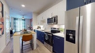 This single-floor residence showcases a thoughtfully designed interior with a contemporary aesthetic throughout. The kitchen is a standout feature, combining navy blue lower cabinetry with crisp white upper cabinets, white quartz countertops, and a geometric-patterned tile backsplash. A full suite of stainless steel appliances—including a French door refrigerator, electric range, microwave, and dishwasher—completes the space. A compact breakfast bar with a butcher block top and seating provides a casual dining option directly adjacent to the kitchen.

The living area is defined by a striking terracotta accent wall adorned with large-scale framed artwork, creating a warm and gallery-like atmosphere. Large windows dressed with patterned curtains invite natural light, and light wood-style flooring runs throughout the main living spaces, adding continuity and warmth.

The bedroom features a deep charcoal accent wall that lends a moody, restful ambiance, complemented by plush carpeting underfoot. The bathroom offers a clean, minimalist design with white shaker-style cabinetry, a single-basin vanity, and light wood flooring, with a passage leading directly into a walk-in closet. Recessed lighting and ceiling fans are present throughout, contributing to a polished, move-in-ready finish.