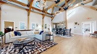 This impressive single-floor space is defined by its dramatic architectural centerpiece: soaring vaulted ceilings supported by exposed timber A-frame trusses with bold black steel bracket hardware and suspended chandelier lighting. Wide-plank hardwood flooring flows seamlessly throughout the main living areas, anchoring the open-concept great room that encompasses generous seating, a dining area with a large dark-wood table and chair seating for a crowd, and a stylish wet bar finished with floor-to-ceiling teal subway tile, white cabinetry, granite countertops, open shelving, and dual beverage refrigerators. Clerestory windows and multiple standard windows bathe the main hall in abundant natural light, enhancing the airy, loft-like atmosphere. A dedicated fitness room offers rubber flooring and a full complement of commercial-grade cardio and strength equipment, with natural light provided by a row of windows along one wall. The game room features hardwood floors, a full-size billiard table, ping-pong table, and casual seating. A bright private office includes hardwood floors, multiple windows, and ample built-in storage. An additional workroom provides carpet flooring, multi-monitor workstations, and open shelving. A large secondary dining room accommodates extensive seating at natural wood communal tables, ideal for group gatherings.
