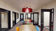 This single-floor collaborative workspace combines open communal areas with enclosed private rooms to accommodate a variety of working styles. The main area features multiple light wood tables paired with an eclectic mix of brightly colored Eames-style chairs in red, blue, yellow, green, and pink, creating an energetic and welcoming atmosphere. A bold red sculptural pendant light serves as a striking centerpiece overhead, drawing the eye upward and adding a dramatic design element to the space. The flooring transitions between smooth grey wood-look planks in the open areas and vibrant patterned carpet in the private rooms, adding visual interest throughout. Black-framed glass doors and interior windows line the perimeter, offering transparency while maintaining acoustic separation between spaces. The private rooms are compact and focused, each equipped with a built-in desk and a single chair, ideal for individual work or small meetings. One room features a textured accent wall, adding warmth and dimension. The overall design balances openness with privacy, modern aesthetics with functional flexibility, making it well-suited for coworking, study, or professional collaboration.