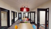 This single-floor collaborative workspace combines open communal areas with enclosed private rooms to accommodate a variety of working styles. The main area features multiple light wood tables paired with an eclectic mix of brightly colored Eames-style chairs in red, blue, yellow, green, and pink, creating an energetic and welcoming atmosphere. A bold red sculptural pendant light serves as a striking centerpiece overhead, drawing the eye upward and adding a dramatic design element to the space. The flooring transitions between smooth grey wood-look planks in the open areas and vibrant patterned carpet in the private rooms, adding visual interest throughout. Black-framed glass doors and interior windows line the perimeter, offering transparency while maintaining acoustic separation between spaces. The private rooms are compact and focused, each equipped with a built-in desk and a single chair, ideal for individual work or small meetings. One room features a textured accent wall, adding warmth and dimension. The overall design balances openness with privacy, modern aesthetics with functional flexibility, making it well-suited for coworking, study, or professional collaboration.