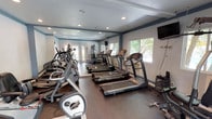This fitness center is designed to provide a comprehensive workout experience within a single, thoughtfully arranged space. The room is lined with full-length mirrors along one wall, which visually expand the area and allow users to monitor their form during exercise. Recessed lighting in the ceiling ensures consistent, even illumination throughout the space, while multiple windows along the exterior walls invite natural light and offer views of the surrounding greenery, including mature palm trees that add a pleasant outdoor connection.

The cardio section is well-stocked with a row of treadmills — including Vision Fitness models — as well as elliptical machines positioned for easy access and comfortable spacing between equipment. A cable and weight machine is also visible toward the rear of the room, catering to strength training needs. A wall-mounted flat-screen television provides entertainment for users during longer cardio sessions.

The flooring is a dark, durable material suited for high-traffic athletic use, and the walls are painted in a clean combination of soft blue and white, lending the space a fresh and motivating atmosphere. A glass-paneled entry door enhances the open feel of the facility. Overall, this fitness center is a functional, well-maintained space suited for a variety of exercise routines.