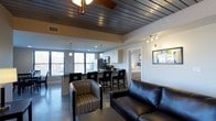 This single-floor space showcases a thoughtfully designed open-concept layout that seamlessly connects the living, dining, and kitchen areas. The striking dark wood plank ceiling serves as a bold architectural focal point, complemented by polished concrete floors that lend an industrial-modern aesthetic throughout the main living area.

The living area is furnished with a sleek black leather sofa and a patterned accent chair, creating a comfortable yet contemporary seating arrangement. A ceiling fan is centrally positioned, enhancing both function and style.

The kitchen features white upper cabinetry, stainless steel appliances including a microwave and refrigerator, and a dark breakfast bar with tall bar seating for multiple guests. Pendant lighting and track lighting illuminate the kitchen and surrounding areas effectively.

Large windows along one wall flood the space with abundant natural light and offer views of surrounding greenery, creating an open and airy atmosphere despite the bold, dark design elements. A glimpse of a bedroom with a neatly made bed is visible through a doorway, suggesting well-defined private quarters adjacent to the main living space. A framed artwork piece adds a decorative touch to the white walls.