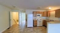 This two-story residence offers a straightforward, functional layout designed for everyday living. The kitchen is a central feature of the main level, equipped with white appliances including a refrigerator, electric range, and over-the-range microwave. Medium-toned wood cabinetry provides ample storage, complemented by countertops in a neutral finish. The kitchen flows naturally into an adjacent hallway, which connects to additional rooms and a staircase leading to the second floor, suggesting a well-organized floor plan. The flooring throughout the main level features a tile-style surface in muted, earthy tones that is both practical and easy to maintain. Ceiling-mounted light fixtures provide consistent overhead lighting throughout. The walls are finished in a clean, neutral white, contributing to a bright and straightforward atmosphere. A fire extinguisher is visibly mounted near the staircase area, reflecting attention to safety. The open sightlines from the kitchen through the hallway enhance the sense of space and connectivity between areas, making the layout feel accessible and easy to navigate across both floors.
