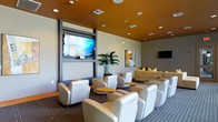 This single-floor space is designed as a stylish communal lounge or media room, offering a comfortable and contemporary environment suited for gatherings or casual viewing. The room is anchored by a large wall-mounted flat-screen television set within a sleek, dark-framed panel, complemented by a secondary display mounted on the adjacent wall, ensuring clear sightlines from multiple seating positions. Rows of modern white leather armchairs are arranged in a theater-inspired layout, each paired with small round side tables finished in warm wood tones, providing convenient surfaces for personal items. The ceiling is a defining architectural feature, finished in a bold amber-orange tone that adds warmth and visual depth to the space, while recessed lighting provides even, ambient illumination throughout. Neutral gray walls create a balanced backdrop that allows the furnishings and ceiling to stand out. A tall, lush indoor plant introduces a natural element near the main display wall. The flooring consists of patterned carpet in muted tones, contributing to the room's acoustic comfort. Accent pillows in yellow tones on a background sofa add subtle pops of color, enhancing the overall inviting ambiance.