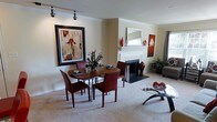 This single-floor space offers an open, cohesive layout that seamlessly connects the dining area and living room, creating a comfortable and inviting atmosphere. The dining area features a rich wood table surrounded by deep red leather chairs, anchored beneath track lighting mounted on the ceiling that provides focused, adjustable illumination. A large, boldly framed piece of abstract wall art adds a dramatic focal point to the dining space.

The living room centers around a classic white-mantled fireplace with a dark surround and raised hearth, decorated with ceramic vases and a rectangular mirror above the mantle. Warm neutral walls and crown molding throughout lend an air of refinement to the space. Plush carpeting in a light neutral tone runs throughout, contributing to the warm and relaxed ambiance.

Accent décor in deep reds and earthy tones ties the rooms together, complementing the bold artwork displayed on the walls. A glass-top coffee table with a sculptural base adds a contemporary touch to the living area, which also benefits from natural light filtering through draped windows. Lush potted greenery further enhances the welcoming character of the space.