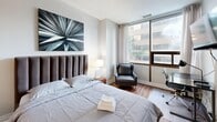 This efficiently designed one-bedroom, one-bathroom unit offers a well-organized open-plan layout that maximizes every square foot. Entry is through a tiled hallway, which provides a clear sightline into the living space and kitchen beyond. The kitchen is positioned at the front of the unit and features white cabinetry, dark granite-style countertops, stainless steel appliances including a full-size refrigerator and electric range, and a peninsula with bar seating. A gold-toned multi-bulb pendant light adds a contemporary accent overhead. The kitchen transitions seamlessly into the bedroom area, where warm hardwood flooring takes over from the tile. Large floor-to-ceiling windows along the exterior wall allow abundant natural light and offer urban views of the surrounding cityscape. The bedroom accommodates a full-size bed with an upholstered channeled headboard, a leather accent chair, and a workspace, making it a functional multi-purpose area. The bathroom is accessed via a folding door and includes white tile flooring, a combined tub and shower, a toilet, and a vanity with dark stone countertops. Throughout, the unit maintains a clean, neutral palette with modern fixtures that contribute to a cohesive and practical aesthetic.