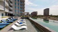 Located at 2902 Carlisle St in Dallas's vibrant Uptown neighborhood, this rooftop amenity level delivers a resort-caliber outdoor experience with commanding views of the surrounding cityscape. The centerpiece is a striking infinity-edge pool that stretches the length of the deck, its crystal-clear water reflecting the Dallas skyline and neighboring architecture. A dedicated tanning shelf lines one side of the pool, fitted with sleek white chaise lounges that invite sun-soaking throughout the day.

Adjacent to the pool, a generously sized lounge area offers plush sectional seating, accent chairs, and a woven rug atop wood-panel decking, creating an inviting space for relaxation or social gatherings. Raised planters filled with ornamental grasses and agave add structured greenery throughout the terrace, complementing the clean, contemporary design aesthetic.

The outdoor kitchen station is equipped with a stainless steel gas grill, built-in sink, and marble-style countertop, all sheltered beneath a pergola and paired with a mounted flat-screen television—ideal for alfresco entertaining. Large-format porcelain tile pavers and glass balustrades carry throughout, maintaining an open, upscale feel. The elevated vantage point provides unobstructed panoramic views of the Dallas urban landscape in every direction.