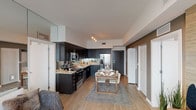 This single-floor residence offers a thoughtfully designed open-concept layout that seamlessly connects the kitchen, dining, and living areas. The kitchen is a standout feature, showcasing dark espresso cabinetry paired with granite countertops, mirrored backsplash, and a full suite of stainless steel appliances, including a french-door refrigerator and built-in microwave. Track lighting overhead illuminates the space, which flows naturally into the dining area, accommodating a wooden table with modern seating.

The living room is bright and welcoming, anchored by a wall-mounted entertainment unit and floor-to-ceiling sliding glass doors that open to a private balcony with expansive waterfront views. Light wood flooring runs throughout the main living areas, enhancing the airy feel.

The primary bedroom features a large mirror panel accent wall, generous natural light through sheer-curtained windows, and wood-look flooring. A secondary bedroom is configured as a versatile children's room with built-in shelving and a dedicated study nook. An additional bedroom offers a quiet, light-filled retreat.

Both bathrooms are finished with granite countertops, dark wood vanities, large-format tile surrounds, and tub-shower combinations, maintaining a clean, contemporary aesthetic throughout.