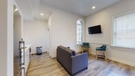 Located at 78 Dorrance St in downtown Providence, Rhode Island, this single-floor unit blends contemporary design with classic architectural details. The living and lounge area features crisp white walls, wood-look flooring, and elegant arched windows that allow generous natural light to fill the space. A wall-mounted television and teal upholstered seating chairs complement the grey sofa, creating a versatile and comfortable gathering area.

The compact kitchenette is efficiently designed with white cabinetry extending to the ceiling, stainless steel appliances including a refrigerator and microwave, and a clean countertop workspace with a sink. The open layout connects the kitchen seamlessly to the living space.

The bedroom is a standout feature, highlighted by a bold herringbone-patterned accent wall in a muted green-grey tone that adds depth and character. Large, multi-pane windows flood the room with natural light, and a wall-mounted television adds convenience. Wood-look flooring continues throughout.

The bathroom is finished with warm-toned large-format floor and wall tiles, offering a clean, modern aesthetic. The overall design of the unit is polished and functional, reflecting contemporary urban living in the heart of Providence.