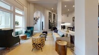 Located at 475 Seven Farms Drive on Daniel Island in Charleston, South Carolina, this single-floor space presents a refined, hospitality-inspired environment characterized by architectural elegance and thoughtful interior design. The entrance corridor immediately establishes the tone, featuring herringbone-patterned hardwood flooring, a dramatic dark-painted coffered ceiling, and arched passageways that draw visitors through the space. Botanical-patterned wallcoverings, a brass wall sconce, and curated artwork add warmth and personality to the arrival experience.

The main lounge area boasts soaring ceilings adorned with exposed dark beams and a crystal chandelier, complemented by floor-to-ceiling windows that flood the space with natural light and offer views of the surrounding greenery. Seating arrangements featuring velvet wingback chairs, rattan accent pieces, and layered rugs create inviting conversation areas. Arched openings and steel-framed mirror panels provide architectural punctuation throughout.

The fully equipped kitchen stands out with deep navy cabinetry, marble-look countertops, open walnut shelving, and a bold tropical palm leaf mural that extends to the ceiling, anchored by statement brass pendant lighting and travertine tile flooring. Together, these elements create a cohesive, upscale atmosphere suited for community gatherings and events.