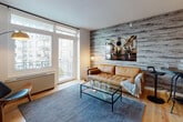 Located at 1277 E 14th St in the Flatbush neighborhood of Brooklyn, NY, this single-floor residence blends modern design with warm, inviting finishes. The living room is a standout space, featuring a striking wood-paneled accent wall with a weathered gray finish, light hardwood flooring, and large floor-to-ceiling glass doors that open to a private balcony, filling the room with natural light and offering views of the surrounding neighborhood greenery. The bedroom continues the warm aesthetic with a bold burnt-orange accent wall, light hardwood floors, and a generously sized window that frames outdoor views. A built-in closet with sliding doors provides ample storage. The kitchen is outfitted with wood-toned cabinetry, stainless steel appliances including a gas range and French door refrigerator, white quartz countertops, and a textured wood-look accent wall that adds depth and character. The bathroom features large-format wall tiles, a wood-vanity with a white countertop, and distinctive navy penny-tile flooring, creating a clean yet visually interesting space. Throughout the residence, cohesive materials and a contemporary design sensibility create a polished, functional living environment.
