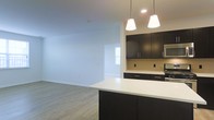 This single-floor space showcases a thoughtfully designed open-concept layout that seamlessly connects the kitchen and living areas. The kitchen is a standout feature, equipped with dark espresso cabinetry that extends to the upper walls, providing ample storage. A crisp white countertop peninsula serves as both a workspace and a casual dining or entertaining surface, offering a striking contrast against the dark cabinetry below. Two stylish pendant lights hang above the peninsula, casting warm, focused illumination over the workspace. Additional recessed lighting enhances the overall brightness of the area. The kitchen also includes stainless steel appliances, including a built-in microwave, adding a modern touch. Light wood-look flooring runs throughout, contributing to a clean and cohesive aesthetic. The open living area beyond the kitchen is bright and airy, with neutral white walls and access to natural light through a window visible in the adjacent room. The overall design emphasizes a contemporary, functional aesthetic, with a neutral color palette that creates a welcoming and versatile environment suitable for a variety of furnishing styles.