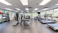 Located at 21 Garrison View Rd in Owings Mills, Maryland, this fitness center offers a thoughtfully designed exercise room that provides ample space for a comprehensive workout experience. The room features durable rubber flooring throughout, ideal for supporting heavy equipment and high-impact activity, while a drop ceiling with recessed panel lighting ensures the space is consistently and evenly lit.

Large multi-pane windows line the walls, inviting natural light and providing pleasant outdoor views of the surrounding greenery, creating an open and energizing atmosphere. The room is well-stocked with a diverse selection of Life Fitness strength machines, including cable systems, leg press, and weight benches, catering to a full range of resistance training needs. A dedicated dumbbell rack offers free weight options, while stability balls and additional accessories support flexibility and functional fitness routines.

The cardio section is equipped with multiple treadmills and an elliptical machine, each positioned near the windows and accompanied by a wall-mounted television for entertainment during workouts. The facility also includes a hand sanitizer station and clearly marked exit signage, reflecting a commitment to cleanliness and safety. French doors provide a welcoming entry point, completing this clean, functional, and well-appointed fitness facility.