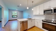 Located at 9 Mill St, this well-appointed single-floor unit showcases a clean, modern design with warm hardwood flooring running throughout the entire space. The kitchen serves as a central highlight, featuring crisp white shaker-style cabinetry extending to the ceiling for ample storage, marble-look countertops, and a full suite of stainless steel appliances including a refrigerator, range with oven, built-in microwave, and dishwasher. Pendant lighting hangs above the L-shaped counter peninsula, which includes an undermount sink, adding both function and style to the space. A large multi-pane window in the adjacent living area allows generous natural light to filter in, offering views of lush greenery outside. The bedrooms are comfortably sized, each featuring the same warm hardwood flooring, neutral light-gray walls, and grid-pane windows that continue the natural light theme. A double-door closet provides substantial storage in one of the rooms. The hallway connects the living spaces to a bathroom finished with white cabinetry, a vanity with mirror, and tile flooring. Recessed lighting and a consistent neutral color palette throughout the unit create a bright, cohesive, and contemporary living environment.