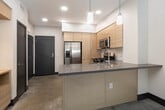 Located at 1800 E Gene Autry Way in Anaheim, California, this single-floor unit showcases a clean, modern design with polished concrete flooring throughout, creating a cohesive and contemporary aesthetic. The open-concept kitchen is well-appointed with light wood cabinetry extending to the ceiling for ample storage, dark charcoal countertops, and a full suite of stainless steel appliances including a gas range, over-the-range microwave, French door refrigerator, and dishwasher. A peninsula with pendant lighting bridges the kitchen to the living area, enhancing the open flow of the space. The living area benefits from generous natural light streaming through large windows fitted with horizontal blinds, and is equipped with a ceiling fan for added comfort. A glass-paneled door provides views of the surrounding greenery. The bathroom features a vanity with wood cabinetry and dark countertop, a tub-shower combination, and accessibility grab bars. Directly accessible from the bathroom is a dedicated walk-in closet with built-in shelving and rail systems, offering practical and organized storage. The overall design is minimalist and functional, suited for modern urban living in the heart of Anaheim.