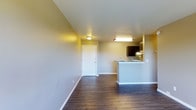 This single-floor space offers a practical and well-organized open-concept layout that seamlessly connects the living area to the kitchen. The living space is defined by rich, dark hardwood flooring that runs throughout, providing a warm contrast against the neutral, light-toned walls. Clean white baseboards frame the room and add a crisp finishing touch to the interior. The kitchen features a peninsula-style breakfast bar with a granite or stone-look countertop, providing both a functional workspace and a casual dining or seating area. Dark espresso-toned upper cabinetry adds depth and a modern aesthetic to the kitchen zone, while overhead lighting ensures the space remains bright and functional. A dedicated flush-mount ceiling light anchors the living area, and additional lighting above the kitchen enhances visibility throughout. The front entry door is centrally positioned along the far wall, offering a straightforward and welcoming access point into the unit. The overall ambiance is clean, contemporary, and move-in ready, with a neutral color palette that provides a versatile backdrop for a variety of décor styles. The open layout maximizes usable space and promotes a comfortable, connected flow between living and cooking areas.