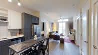 This single-floor residence showcases a thoughtfully designed open-concept layout that seamlessly connects the kitchen, dining, and living areas into one cohesive and functional space. The kitchen is outfitted with sleek gray cabinetry, stainless steel appliances including a French door refrigerator, and a striking marble-topped breakfast bar with seating — ideal for casual dining and entertaining. Light wood-tone flooring runs throughout, lending warmth and continuity to the space. The dining area is positioned adjacent to the kitchen, offering a comfortable transition into the expansive living room beyond. The living room is bright and welcoming, anchored by a ceiling fan and bathed in natural light streaming through large floor-to-ceiling windows and a sliding glass door that opens to a private balcony. The outdoor access enhances the sense of openness and connects the interior to the outdoors. A glimpse into the bedroom reveals a well-proportioned sleeping space with ample room for furnishings. Overall, the residence presents a clean, contemporary aesthetic with a balanced mix of functional design and modern finishes, making it a comfortable and stylish place to call home.