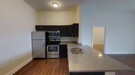 This single-floor space offers a thoughtfully designed layout that balances functionality and modern style. The kitchen serves as a central highlight, showcasing rich dark cabinetry that provides ample storage and creates a striking contrast against the granite countertops. Stainless steel appliances — including a full-size refrigerator, electric range, built-in microwave, and a stainless steel undermount sink — equip the kitchen with everything needed for everyday living. A kitchen island or peninsula extends the counter space and adds versatility for casual dining or meal preparation. The warm hardwood-style flooring runs throughout the kitchen and adjoining areas, adding a cohesive and inviting character to the space. The open sightlines from the kitchen connect seamlessly to the surrounding living areas, creating an airy and interconnected feel. Neutral wall tones complement the overall design, lending a clean and move-in-ready aesthetic. A doorway visible from the kitchen hints at additional rooms beyond, suggesting a practical and well-organized floor plan. The space is well-lit with ceiling-mounted fixtures, ensuring a bright and welcoming atmosphere throughout.