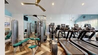 The fitness center at 200 Crowne Club Dr in Winston-Salem, NC, offers a thoughtfully designed exercise space that balances functionality with an inviting aesthetic. The room features striking vaulted ceilings that lend an open, airy feel, painted in crisp white and accented by soft sky-blue walls that create a calm, motivating atmosphere. Underfoot, a distinctive herringbone-patterned wood-look flooring adds visual interest and a polished finish throughout the space. Large windows along one wall invite abundant natural light and provide views of the outdoor surroundings, including what appears to be a pool area, enhancing the workout experience. Full-length wall mirrors amplify the sense of space while allowing users to monitor their form. The facility is well-equipped with a row of treadmills and elliptical trainers for cardiovascular workouts, alongside a selection of strength-training machines, a cable machine, a weight bench, and a variety of free weights. Ceiling fans and recessed lighting ensure comfort and visibility at any time of day. A mounted flat-screen television adds an entertainment option for users. The room is accessed through charming French doors with glass panes, adding an elegant architectural touch. Overall, the fitness center provides a comprehensive and welcoming environment for residents to pursue their health and wellness goals.