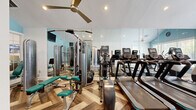 The fitness center at 200 Crowne Club Dr in Winston-Salem, NC, offers a thoughtfully designed exercise space that balances functionality with an inviting aesthetic. The room features striking vaulted ceilings that lend an open, airy feel, painted in crisp white and accented by soft sky-blue walls that create a calm, motivating atmosphere. Underfoot, a distinctive herringbone-patterned wood-look flooring adds visual interest and a polished finish throughout the space. Large windows along one wall invite abundant natural light and provide views of the outdoor surroundings, including what appears to be a pool area, enhancing the workout experience. Full-length wall mirrors amplify the sense of space while allowing users to monitor their form. The facility is well-equipped with a row of treadmills and elliptical trainers for cardiovascular workouts, alongside a selection of strength-training machines, a cable machine, a weight bench, and a variety of free weights. Ceiling fans and recessed lighting ensure comfort and visibility at any time of day. A mounted flat-screen television adds an entertainment option for users. The room is accessed through charming French doors with glass panes, adding an elegant architectural touch. Overall, the fitness center provides a comprehensive and welcoming environment for residents to pursue their health and wellness goals.