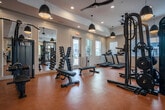 Located at 5060 City Station Dr in Collegedale, Tennessee, this single-floor fitness center is designed to support a comprehensive workout experience across multiple distinct zones. The space is finished with warm terracotta-toned flooring throughout, providing a cohesive and durable surface suited for a variety of exercises.

The main workout area is generously sized and houses an array of strength training equipment, including a multi-function cable machine, adjustable benches, a full dumbbell rack, and free weights such as kettlebells and medicine balls. Ceiling-mounted pendant lights in mixed styles—ranging from black dome fixtures to woven rattan shades—add a distinctive design element alongside recessed lighting and ceiling fans.

A dedicated cardio zone features treadmills, elliptical machines, and a rowing machine, all positioned near windows fitted with white shutters that allow natural light to fill the room. Large mirrors along the walls enhance the sense of openness throughout.

A separate studio room offers a wall-mounted wooden barre, floor-to-ceiling mirrors, a wall-mounted TV, and ample open floor space for stretching or group classes. Additional amenities include a beverage refrigerator, storage shelving, and access to a restroom with brick accent walls.