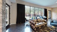This sophisticated single-floor clubroom lounge is designed with a bold contemporary aesthetic that balances comfort and style. The main lounge area is generously sized, anchored by a striking mix of geometric-patterned sofas and sleek blue leather swivel chairs with gold-toned bases, all arranged around low-profile coffee tables atop a graphic monochrome area rug. A row of expansive floor-to-ceiling windows floods the space with natural light and frames verdant outdoor views of mature trees and palms, creating a seamless connection to the surrounding landscape. Dramatic black drapery panels provide contrast and light control. A dedicated billiards area with a full-size pool table serves as a central entertainment feature, complemented by pendant lighting overhead. A textured accent wall with integrated lighting surrounds a wall-mounted flat-screen television, flanked by curated artwork and additional seating. The fully equipped kitchen is outfitted with high-gloss cream cabinetry, striking blue-grey granite countertops, a subway tile backsplash, under-cabinet lighting, and a matte black pull-down faucet. A casual dining nook with bistro-style tables and upholstered chairs is nestled near the windows, ideal for informal gatherings. Recessed lighting throughout ensures a warm, welcoming ambiance across the entire space.