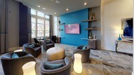 This sophisticated single-floor lounge space is defined by its striking contemporary design and generous proportions. Soaring ceilings create an immediate sense of grandeur upon entry, while floor-to-ceiling windows flood the interior with abundant natural light and offer views of the surrounding neighborhood. The entrance foyer sets an elegant tone, featuring a sleek console table adorned with cobalt blue glass lamps flanking a large abstract artwork, establishing the space's signature color palette of navy, teal, and warm gray.

The main lounge area is thoughtfully arranged with curved, upholstered barrel chairs in charcoal gray accented by teal throw pillows, grouped around a central leather ottoman. Glowing accent lamps add warmth and visual interest throughout. A bold teal feature wall serves as a focal point, hosting a wall-mounted flat-screen television flanked by floating wooden shelves displaying decorative objects.

Separate seating zones include glass-top bistro tables paired with two-tone leather and fabric chairs, ideal for more intimate gatherings. Dark navy cylindrical columns provide architectural punctuation throughout the space. The patterned area rugs ground each seating cluster, contributing to a cohesive, upscale ambiance suited for communal use.