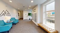 The Vantage – Live Bellingham presents a thoughtfully designed single-floor lobby and corridor space that makes a strong first impression. The entrance foyer features large black-framed glass doors and generous windows that flood the space with natural light, creating an open and airy atmosphere. Plush carpeted flooring in a neutral tone runs throughout, complemented by crisp white walls and recessed lighting that add to the clean, contemporary aesthetic. Tufted leather benches and vibrant teal accent chairs are strategically placed to offer comfortable seating for residents and guests alike, while potted tropical plants introduce a refreshing touch of greenery. A dedicated mail center is built into the wall, providing residents with convenient access to their correspondence. Framed black-and-white photography lines the walls of the extended corridor, lending the space a gallery-like quality as it leads residents deeper into the building. A secure access door with a keypad entry system provides controlled passage to residential areas. The overall ambiance is polished and welcoming, balancing modern design sensibilities with practical residential amenities, making the common areas of The Vantage both functional and visually appealing.