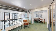 Located at 525 N 7th St in San Jose, California, this thoughtfully designed communal space offers a compelling blend of functionality and modern aesthetics across a single floor. The layout is organized to accommodate both intimate gatherings and collaborative work, featuring distinct zones that serve different purposes while maintaining a cohesive visual identity throughout.

At the heart of the space is a sleek private dining room enclosed by sliding glass-and-dark-frame partitions, centered around a striking solid wood communal table paired with matching wood chairs. A sculptural pendant light hangs above the table, while large windows at the far end flood the room with natural light and offer views of the surrounding urban exterior. Curated wall art adds a refined, gallery-like touch.

Adjacent to the dining area, a relaxed lounge zone features green-toned carpet that lends a warm, organic contrast to the white walls and crisp recessed lighting. Comfortable gray booth seating with sage accent cushions is complemented by leather armchairs and wood side tables, creating inviting nooks for casual conversation or focused work. An exposed metallic ductwork element runs along the upper wall, adding an industrial accent to the otherwise polished interior.

The space also includes what appears to be a private phone booth or pod near the entrance, offering a semi-enclosed option for added privacy. Glass railings and open sightlines contribute to an airy, connected atmosphere throughout.