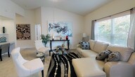 Located at 9821 Chapel Rd in Waco, Texas, this single-story home offers a well-designed layout with a variety of functional and inviting spaces. The living room is a standout area, featuring dramatic vaulted ceilings that lend a sense of openness and volume. Natural light filters in through windows dressed with blinds and drapes, illuminating the neutral-toned walls and plush carpeted floors. A dedicated nook with a built-in desk provides a practical workspace tucked neatly off the main living area. The dining area accommodates comfortable seating and benefits from large windows that bring in outdoor views and additional natural light, complemented by a wall-mounted mirror that enhances the sense of space. The kitchen is equipped with ample cabinetry featuring warm wood accents, white appliances including a refrigerator, range, and microwave, and generous counter space with a double-basin sink. Tile flooring grounds the kitchen and extends into adjacent areas. The bedroom offers carpeted floors, vaulted ceilings, a walk-in closet with built-in shelving, and a window with garden views. Two full bathrooms, each with a tub-shower combination and overhead cabinetry, round out the layout, providing practical and clean utility throughout the home.
