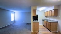 This single-floor residence showcases a practical open-concept design that seamlessly connects the kitchen and living areas. The kitchen is equipped with light wood cabinetry, a coil-top electric range, a built-in dishwasher, and a countertop that extends into a peninsula, creating a natural division between the cooking and living spaces. Wood-style flooring runs throughout the kitchen area, lending warmth and easy maintenance underfoot. The adjacent living area features soft wall-to-wall carpeting, neutral wall tones, and a window that invites natural light into the room, creating a bright and welcoming atmosphere. A textured ceiling spans the entire space, adding subtle dimension overhead. The open flow between the kitchen and living room makes the layout well-suited for everyday living, offering both functional cooking space and a comfortable area to relax. The neutral color palette throughout provides a clean, versatile backdrop that can be easily personalized. Overall, the space is thoughtfully arranged to maximize usability within a straightforward, single-level floor plan.