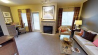 Located at 11400 Olympus Way in Gig Harbor, Washington, this single-floor residence offers a thoughtfully designed living space that balances comfort and style. The living room serves as the heart of the home, centered around a tile-surround fireplace that provides a warm and inviting focal point. A framed landscape artwork is displayed prominently above the mantle, adding a refined decorative touch. Warm yellow walls contribute to the bright and cheerful atmosphere of the room, complemented by rich, patterned drapery in earth tones that frame multiple windows. These windows allow generous natural light to fill the space, creating an open and airy feel. The living area is finished with plush wall-to-wall carpeting underfoot, adding to the overall sense of coziness. Track lighting on the ceiling provides adjustable illumination to enhance the room's functionality. The layout is well-proportioned, offering ample space for seating arrangements that encourage both relaxation and conversation. Overall, this residence presents a warm, comfortable, and tastefully appointed interior well-suited to the serene surroundings of Gig Harbor.
