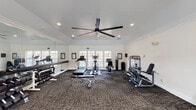 This fitness center in Robertsdale, Alabama offers a thoughtfully designed, single-floor workout facility built to support a wide range of exercise routines. The space features vaulted ceilings accented with crown molding and is finished in crisp white throughout, creating a clean and energizing atmosphere. A large black ceiling fan anchors the main area, ensuring comfortable airflow during workouts.

Natural light pours in through multiple windows and glass-paneled doors along the walls, with views to the outdoors. The main workout area is outfitted with cardio equipment including treadmills, an elliptical trainer, and a stationary bike, all arranged on durable patterned carpet flooring. A TKO strength machine provides resistance training options, while a full dumbbell rack lines one wall alongside a weight bench for versatile strength training.

A floor-to-ceiling mirror runs along one wall, expanding the sense of space and allowing users to monitor their form. A dedicated open area with a floor mat and a medicine ball rack offers space for stretching, functional training, or floor exercises. Recessed lighting complements the natural light, keeping the space bright and welcoming at all hours. Informational boards are posted on the walls, and a water station is conveniently available for users throughout their workout.