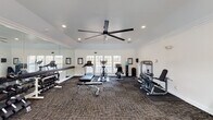 This fitness center in Robertsdale, Alabama offers a thoughtfully designed, single-floor workout facility built to support a wide range of exercise routines. The space features vaulted ceilings accented with crown molding and is finished in crisp white throughout, creating a clean and energizing atmosphere. A large black ceiling fan anchors the main area, ensuring comfortable airflow during workouts.

Natural light pours in through multiple windows and glass-paneled doors along the walls, with views to the outdoors. The main workout area is outfitted with cardio equipment including treadmills, an elliptical trainer, and a stationary bike, all arranged on durable patterned carpet flooring. A TKO strength machine provides resistance training options, while a full dumbbell rack lines one wall alongside a weight bench for versatile strength training.

A floor-to-ceiling mirror runs along one wall, expanding the sense of space and allowing users to monitor their form. A dedicated open area with a floor mat and a medicine ball rack offers space for stretching, functional training, or floor exercises. Recessed lighting complements the natural light, keeping the space bright and welcoming at all hours. Informational boards are posted on the walls, and a water station is conveniently available for users throughout their workout.