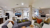 This single-floor communal space is thoughtfully designed to foster comfort and social interaction. The open-concept layout is anchored by striking stone-clad columns that add texture and architectural interest throughout. Recessed lighting and a tray ceiling with decorative molding lend a polished, upscale feel to the interior.

The lounge area features contemporary sectional sofas and accent chairs in neutral and earthy tones, accented with olive green and rust-colored throw pillows that add warmth and personality. Sleek gold accent tables serve as functional yet decorative focal points. A large flat-screen TV display wall creates an entertainment hub, complemented by pendant lighting overhead.

Adjacent to the lounge, a kitchen bar area is equipped with a gas cooktop, white countertops, and bar-height seating with transparent acrylic chairs, offering a clean and modern aesthetic. A secondary seating nook with a high-top table and chairs provides additional gathering space.

Natural light filters in through large windows on one side of the space, brightening the white tile flooring and creating an airy, welcoming ambiance. The overall design balances functionality with contemporary style, making this a versatile and well-appointed shared space.