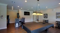 This single-floor space is designed with entertainment and comfort in mind, centered around a well-appointed billiards room that serves as the heart of the layout. The room features rich hardwood flooring that adds warmth and character throughout. A full-size pool table anchors the space, illuminated by an elegant three-shade pendant light fixture suspended from the ceiling by chains, providing focused, ambient lighting over the playing surface. Recessed lighting is distributed evenly across the ceiling, ensuring the entire room is well-lit, while crown molding adds a refined architectural touch along the ceiling's perimeter. A wall-mounted flat-screen television is positioned for easy viewing, complementing the entertainment-focused design. A high-top bar-style seating area with upholstered chairs sits along one wall, ideal for spectators or casual gatherings. French doors introduce natural light and offer a visual connection to an adjacent area. Framed wall art adds a personal, curated touch to the décor. The neutral wall tones paired with dark cabinetry and furniture create a sophisticated yet inviting contrast. Overall, the space strikes a balance between a relaxed recreational environment and polished interior design.