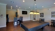 This single-floor space is designed with entertainment and comfort in mind, centered around a well-appointed billiards room that serves as the heart of the layout. The room features rich hardwood flooring that adds warmth and character throughout. A full-size pool table anchors the space, illuminated by an elegant three-shade pendant light fixture suspended from the ceiling by chains, providing focused, ambient lighting over the playing surface. Recessed lighting is distributed evenly across the ceiling, ensuring the entire room is well-lit, while crown molding adds a refined architectural touch along the ceiling's perimeter. A wall-mounted flat-screen television is positioned for easy viewing, complementing the entertainment-focused design. A high-top bar-style seating area with upholstered chairs sits along one wall, ideal for spectators or casual gatherings. French doors introduce natural light and offer a visual connection to an adjacent area. Framed wall art adds a personal, curated touch to the décor. The neutral wall tones paired with dark cabinetry and furniture create a sophisticated yet inviting contrast. Overall, the space strikes a balance between a relaxed recreational environment and polished interior design.