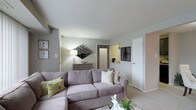 This single-floor residential space offers a well-appointed and comfortable living environment designed with a clean, neutral aesthetic. The living area serves as the heart of the space, featuring plush wall-to-wall carpeting underfoot and smooth, light-toned walls that create a calm and inviting atmosphere. A generous sectional sofa anchors the room, complemented by decorative accent pillows and a coffee table, making the space ideal for relaxing or entertaining. Wall art and a geometric mirror add visual interest and personality to the room, while a console table with a table lamp provides warm, ambient lighting. The layout flows naturally, with doorways leading to additional areas of the home, including what appears to be a kitchen or utility space visible through a pass-through opening. The entry area is clearly defined, offering a functional transition into the main living space. White interior doors and clean trim work contribute to the overall tidy and well-maintained character of the residence. The single-level layout ensures easy accessibility and a cohesive flow throughout, making the space both practical and comfortable for everyday living.