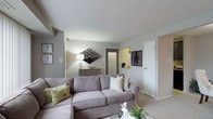 This single-floor residential space offers a well-appointed and comfortable living environment designed with a clean, neutral aesthetic. The living area serves as the heart of the space, featuring plush wall-to-wall carpeting underfoot and smooth, light-toned walls that create a calm and inviting atmosphere. A generous sectional sofa anchors the room, complemented by decorative accent pillows and a coffee table, making the space ideal for relaxing or entertaining. Wall art and a geometric mirror add visual interest and personality to the room, while a console table with a table lamp provides warm, ambient lighting. The layout flows naturally, with doorways leading to additional areas of the home, including what appears to be a kitchen or utility space visible through a pass-through opening. The entry area is clearly defined, offering a functional transition into the main living space. White interior doors and clean trim work contribute to the overall tidy and well-maintained character of the residence. The single-level layout ensures easy accessibility and a cohesive flow throughout, making the space both practical and comfortable for everyday living.
