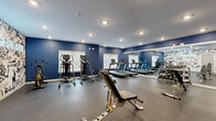 This fitness center offers a thoughtfully designed workout environment spread across a single level, providing ample space for a full range of exercise activities. The interior is defined by bold navy blue walls that create an energetic atmosphere, complemented by a striking graffiti-style mural accent wall that adds an urban, dynamic character to the space.

The facility is outfitted with a comprehensive selection of equipment. The cardio zone features multiple treadmills aligned along a full-length wall mirror, which enhances the sense of depth and space while allowing users to monitor their form. Elliptical trainers and a rowing machine round out the cardio offerings. On the strength training side, the space includes a cable machine with TRX suspension straps, adjustable weight benches, a dumbbell rack, and a dedicated functional training area with a tiered rack of medicine balls and stability balls in various sizes.

Durable rubber flooring covers the entire area, providing both safety and comfort during workouts. Recessed ceiling lighting ensures the space is well-illuminated throughout, while multiple windows fitted with blinds allow for natural light. The overall layout is open and efficiently organized, supporting a variety of individual training styles and fitness routines.