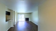 This single-floor space features an open living area defined by rich, dark hardwood-style flooring that reflects natural light streaming in from a large sliding glass door. The door serves as both a functional entryway to the outdoors and a source of abundant natural light, framing a lush green view that adds a sense of connection to the exterior environment. A centrally positioned fireplace with a dark surround serves as a focal point of the room, providing both visual interest and a functional feature for added warmth and ambiance. The walls are finished in a neutral, light tone that complements the overall brightness of the space, while white baseboards offer a clean contrast against the flooring. The textured ceiling adds a subtle architectural detail overhead. The layout is open and unobstructed, allowing for flexible furniture arrangement and a versatile use of the space. Overall, the area presents a straightforward, well-lit environment with practical features, making it a functional and adaptable living space suitable for a variety of needs.
