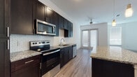 This thoughtfully designed single-floor unit blends modern aesthetics with functional living. The kitchen is a standout feature, showcasing floor-to-ceiling dark espresso cabinetry, granite countertops, and a full suite of stainless steel appliances including a French door refrigerator, electric range, over-the-range microwave, and dishwasher. A geometric hexagonal tile backsplash adds visual interest, while a center island with pendant lighting and additional granite workspace enhances both utility and style. Light wood-tone luxury vinyl plank flooring flows seamlessly throughout the space, creating a cohesive and contemporary feel. The open living area is bright and airy, benefiting from abundant natural light through multiple windows fitted with blinds and a set of French doors that lead to the exterior. A ceiling fan keeps the space comfortable year-round. The bathroom is equally refined, featuring a dual-sink vanity topped with speckled granite, dark cabinetry with brushed nickel hardware, a large mirror, a walk-in glass-enclosed shower, and a spacious walk-in closet. White walls and tile flooring in the bathroom contribute to a clean, modern finish throughout. Overall, the unit presents a polished and move-in-ready living environment.
